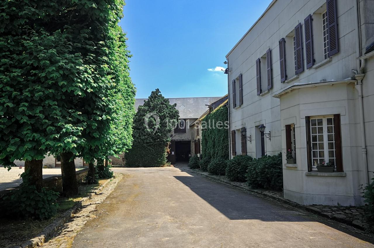 Allée pavée bordée d'arbres et de bâtiments blancs aux volets fermés, sous un ciel bleu dégagé.