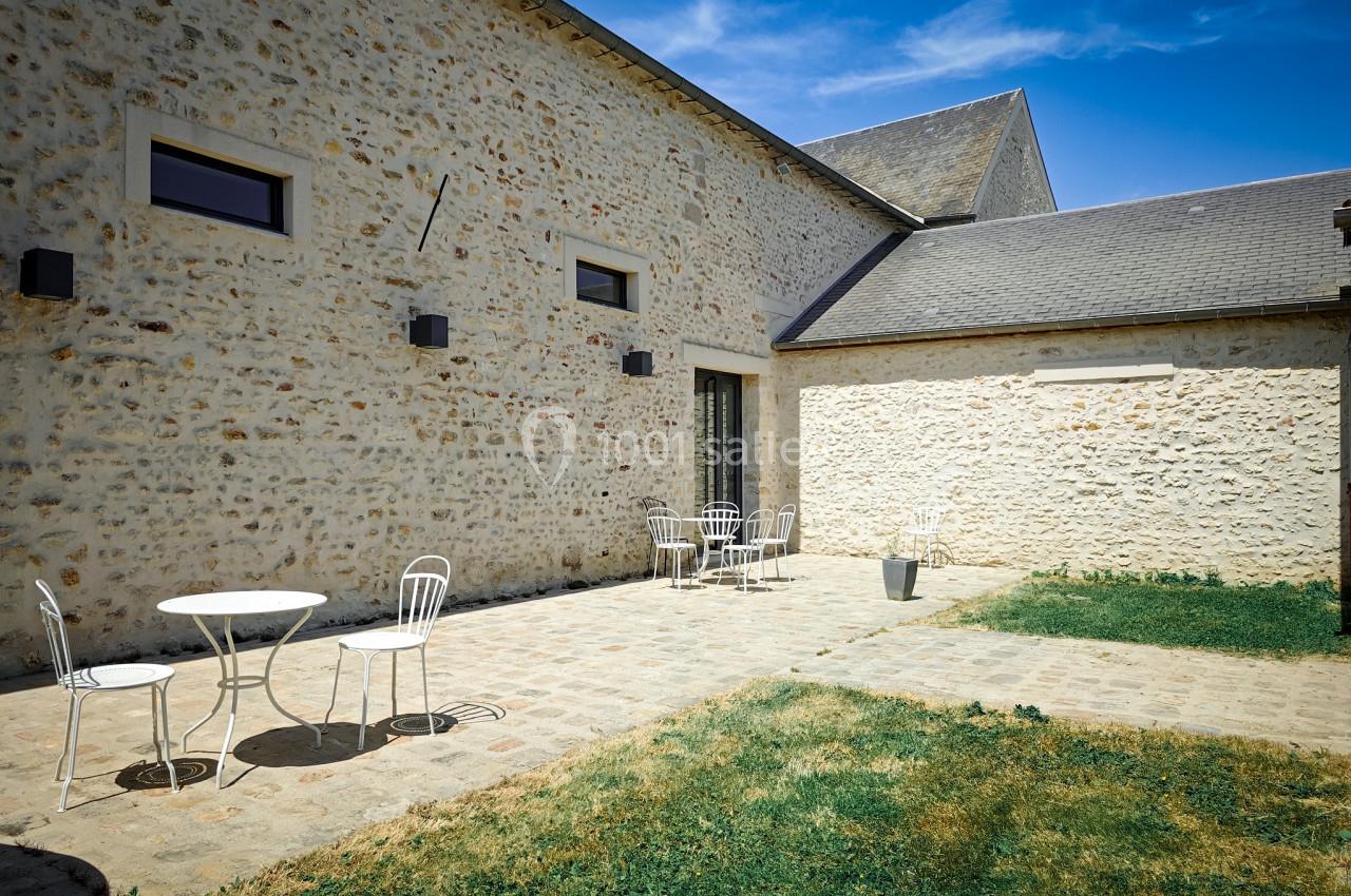 Cour pavée avec des murs en pierre, tables et chaises blanches, partiellement ombragée sous un ciel bleu.
