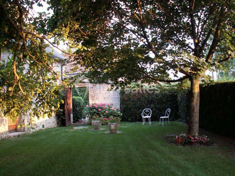 Jardin avec pelouse, arbre ombragé, chaises blanches en métal, pots de fleurs et façade en pierre en arrière-plan.