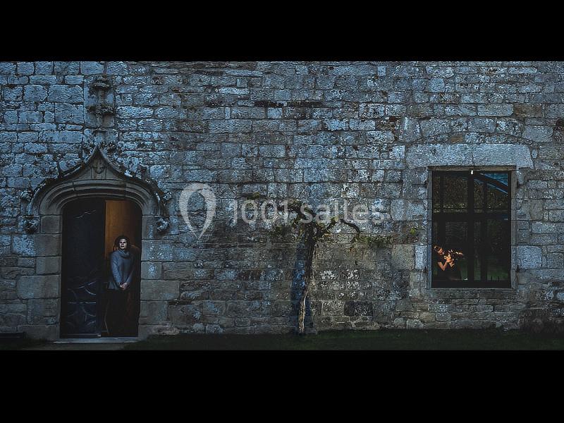 Une femme se tient dans l'encadrement d'une porte ouverte, près d'un mur en pierre avec une fenêtre éclairée.