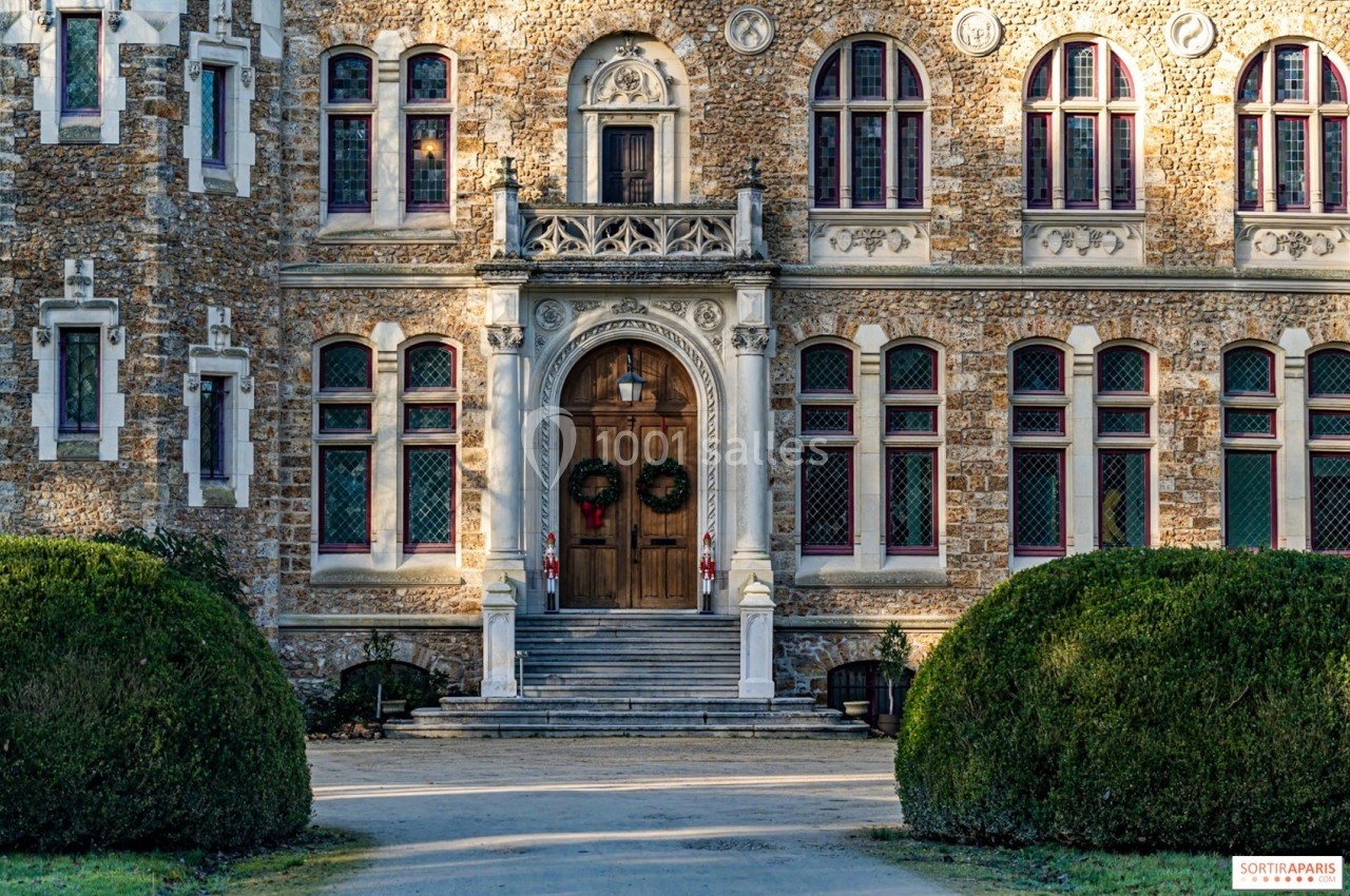 Façade en pierre d'un bâtiment historique avec une porte en bois ornée de couronnes de Noël, entourée de buissons.