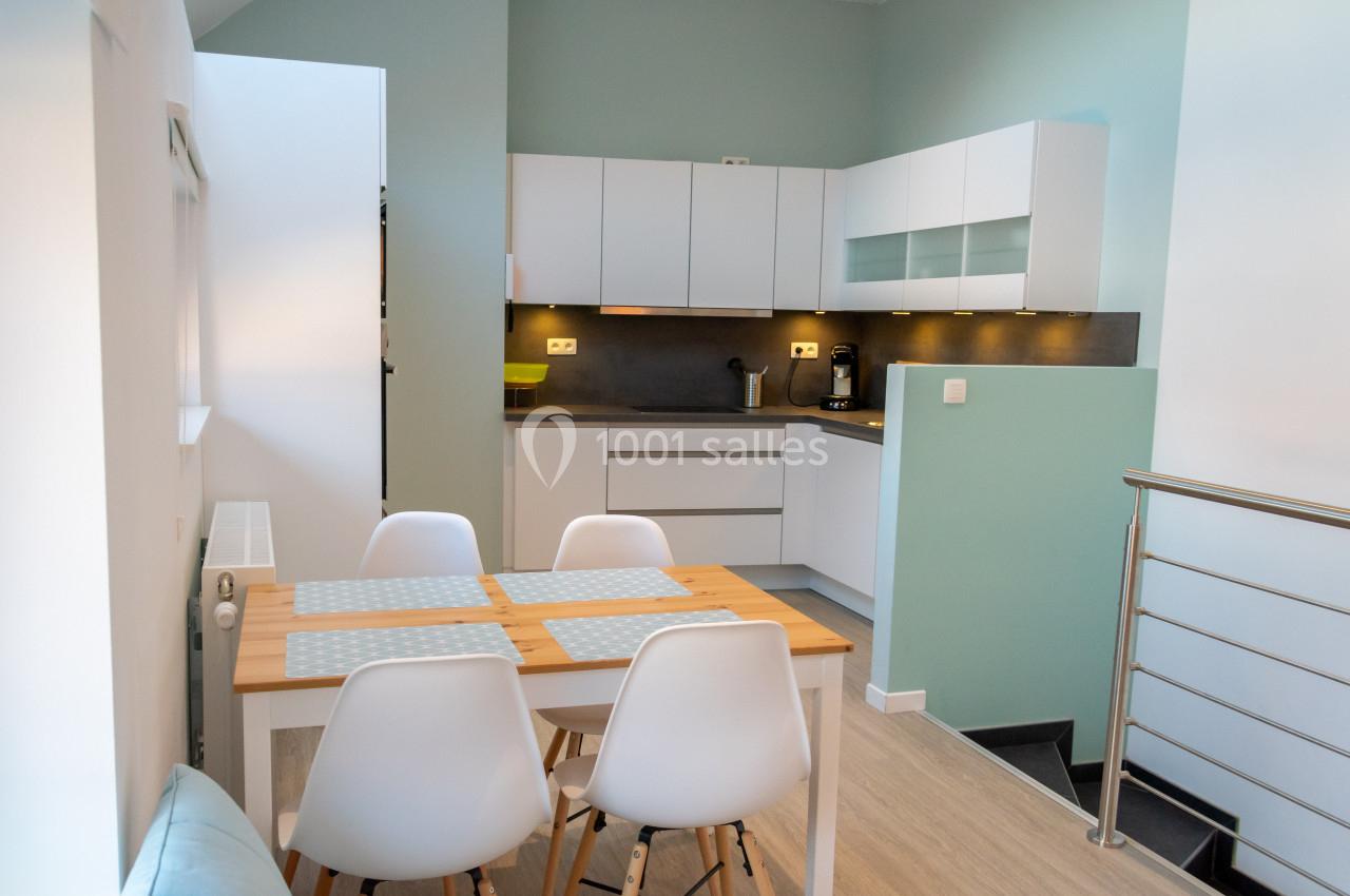 Cuisine moderne avec meubles blancs, table en bois clair et chaises blanches, éclairée par une lumière naturelle.