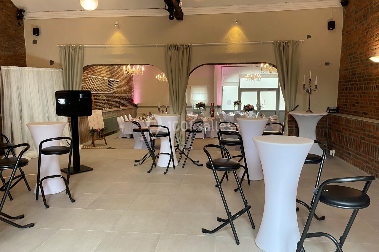 Salle de réception lumineuse avec murs en briques, tables hautes blanches et chaises noires, décorée pour un événement.