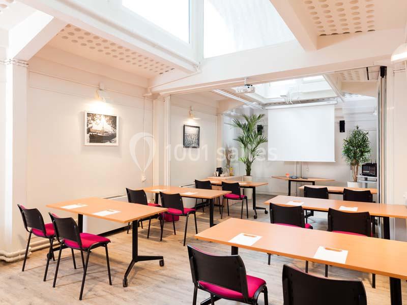 Salle lumineuse aménagée avec des tables en bois, des chaises noires et rouges, un écran de projection et des plantes.