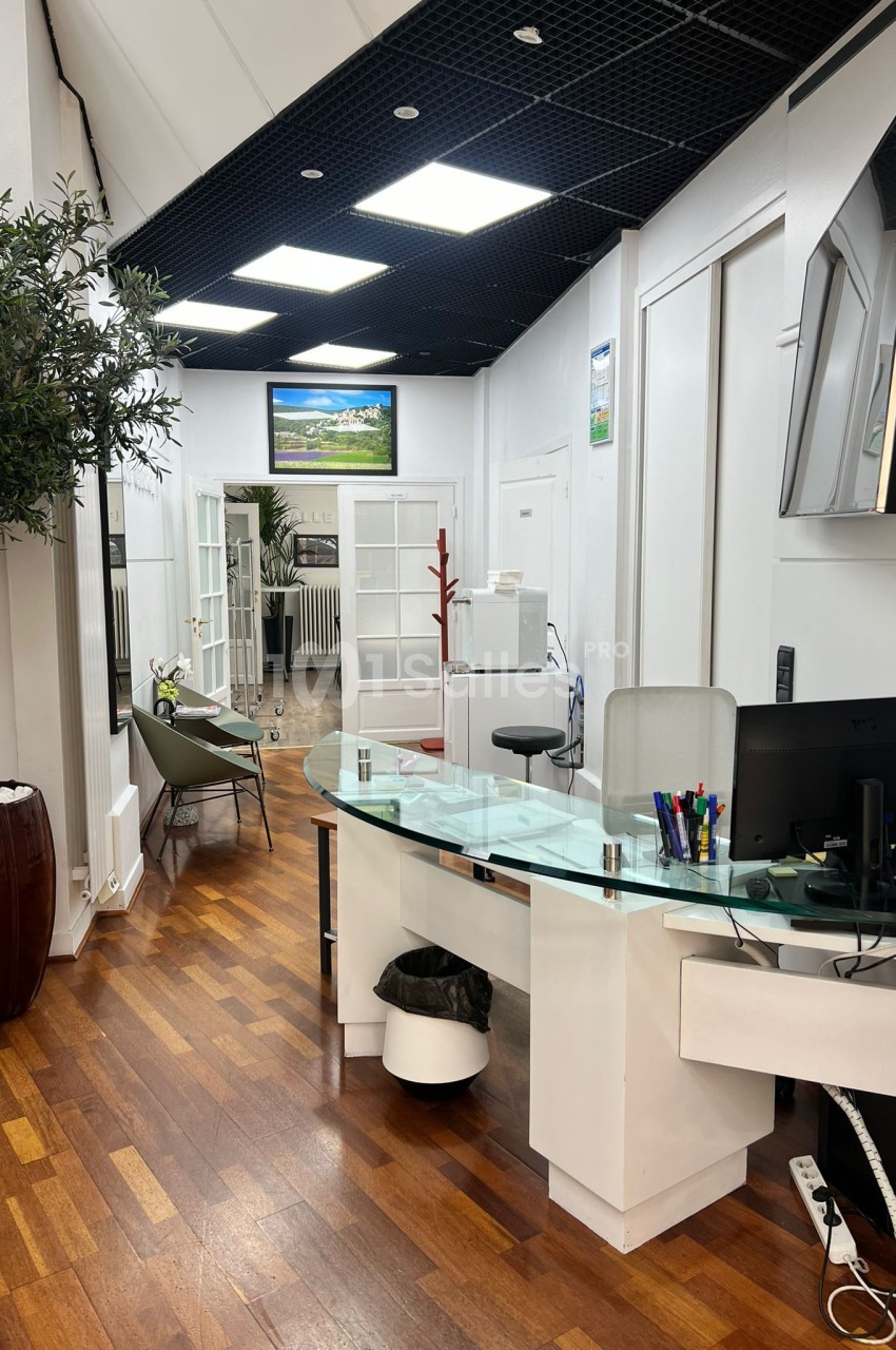 Bureau moderne avec comptoir en verre, mobilier blanc, sol en bois, et plantes décoratives dans un espace lumineux.