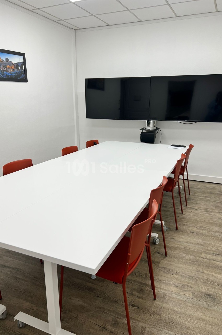 Salle de réunion avec une grande table blanche, des chaises rouges, deux écrans muraux et un sol en bois clair.