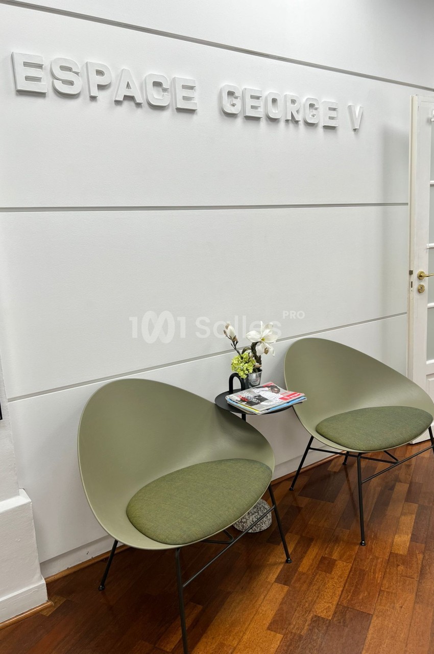 Deux fauteuils verts autour d'une petite table avec des magazines et des fleurs, dans un espace nommé ’Espace George V’.