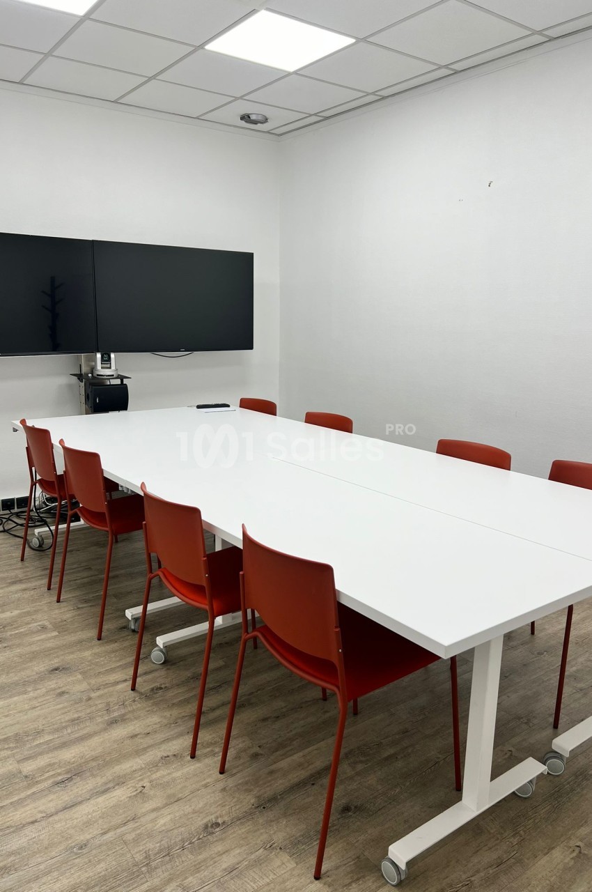 Salle de réunion avec table blanche, chaises rouges, deux écrans muraux et sol en parquet clair.