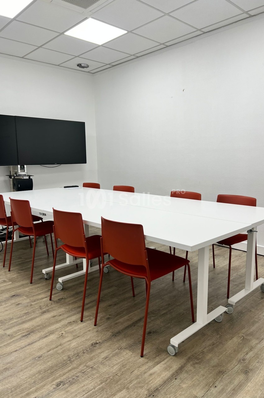 Salle de réunion avec table blanche, chaises rouges, écran mural et sol en parquet clair.
