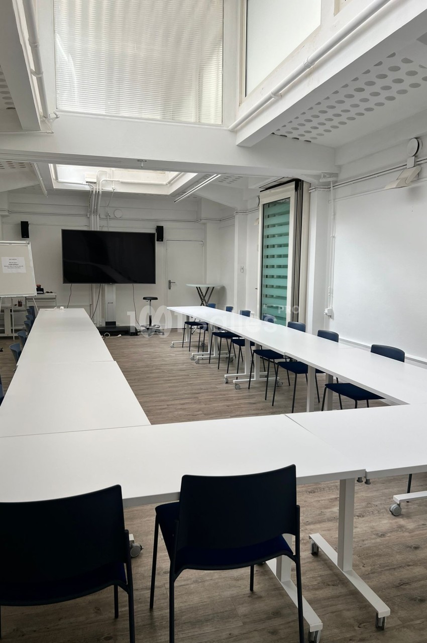 Salle de réunion lumineuse avec tables blanches en disposition en U, chaises noires et écran de présentation.