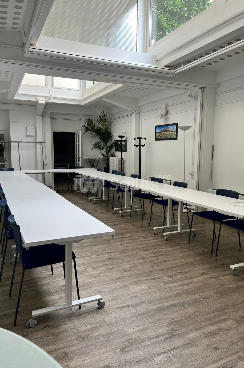 Salle de réunion lumineuse avec de grandes tables blanches disposées en U, chaises bleues et plantes en arrière-plan.