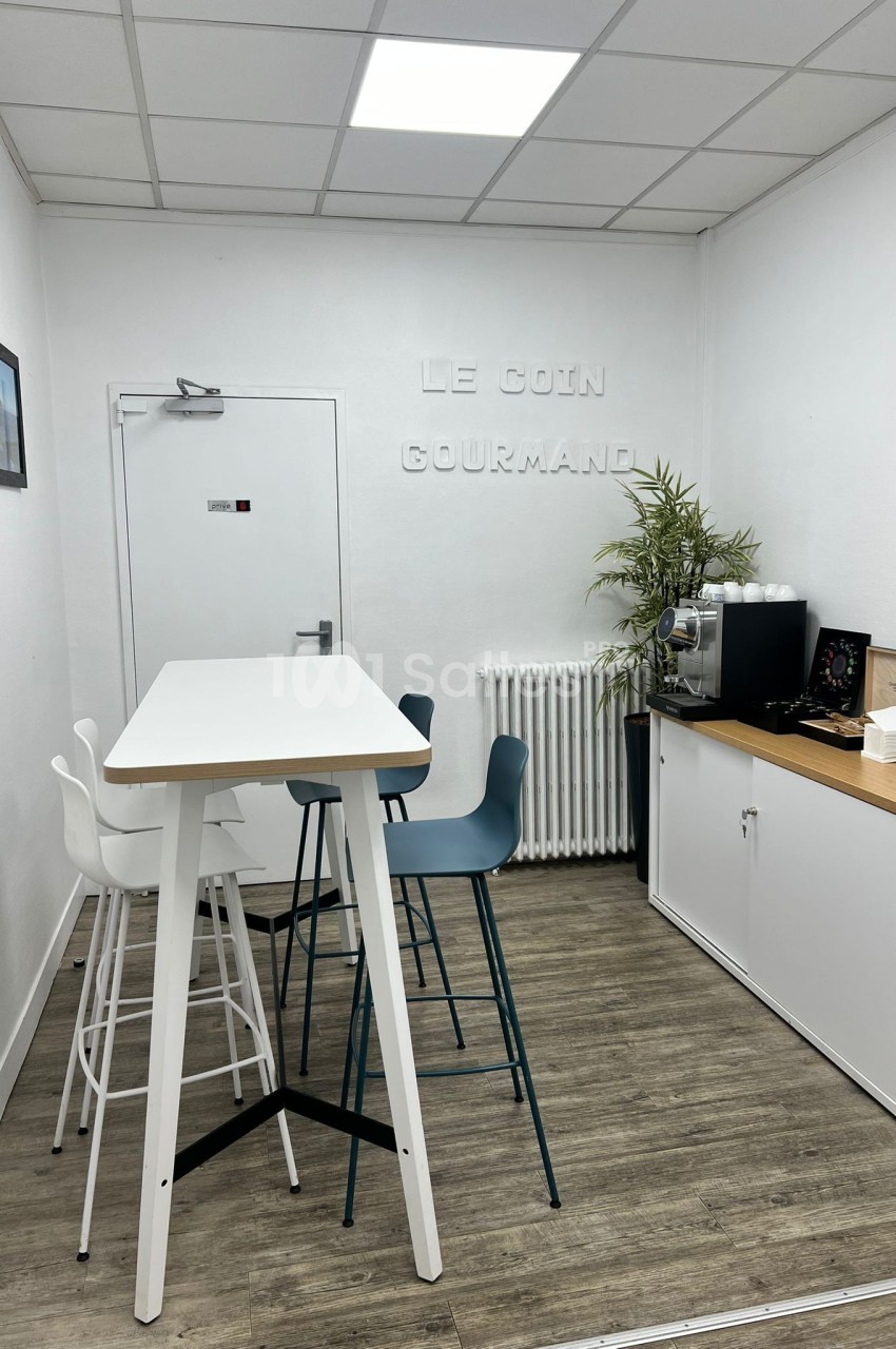 Espace de pause avec table haute, chaises, machine à café, plante et décoration murale dans une pièce lumineuse.