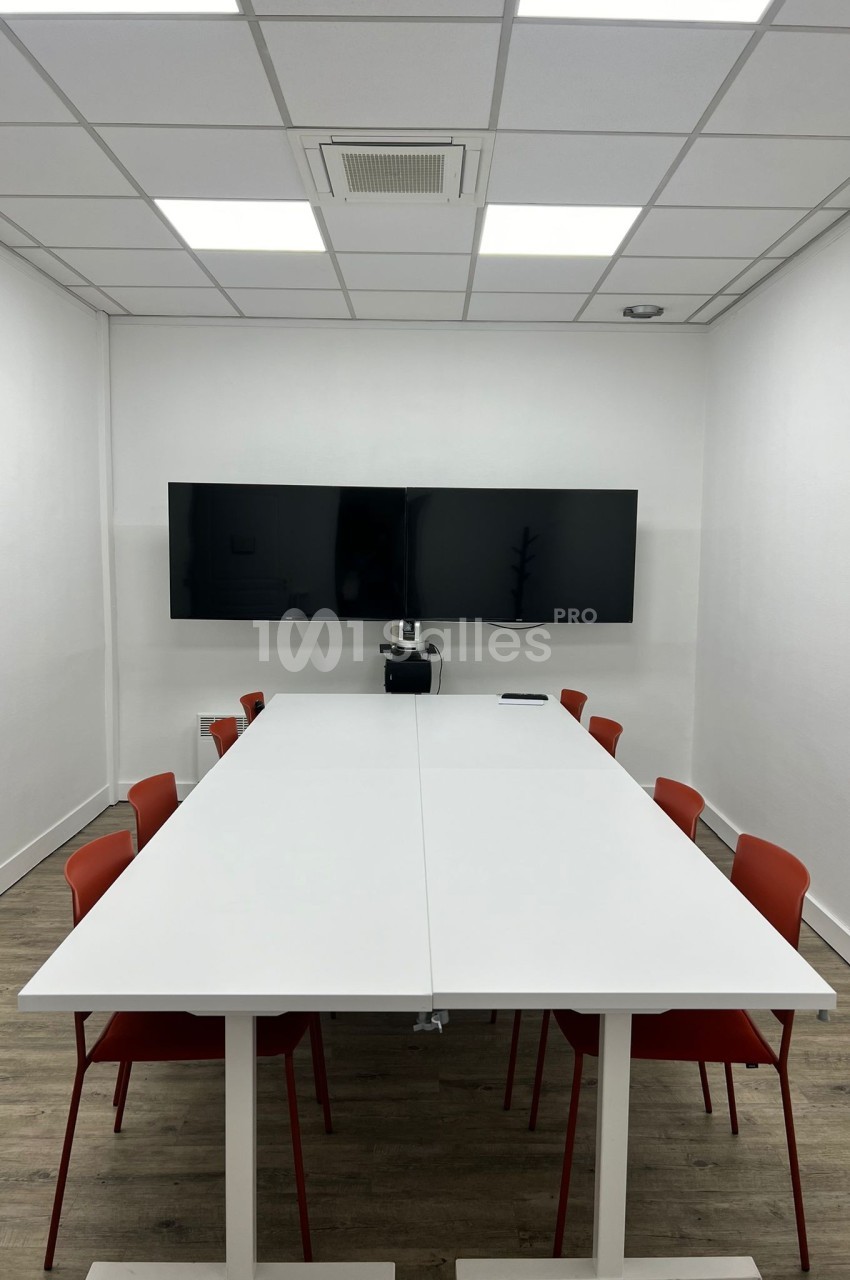 Salle de réunion avec table blanche, chaises rouges, écran mural et éclairage au plafond.