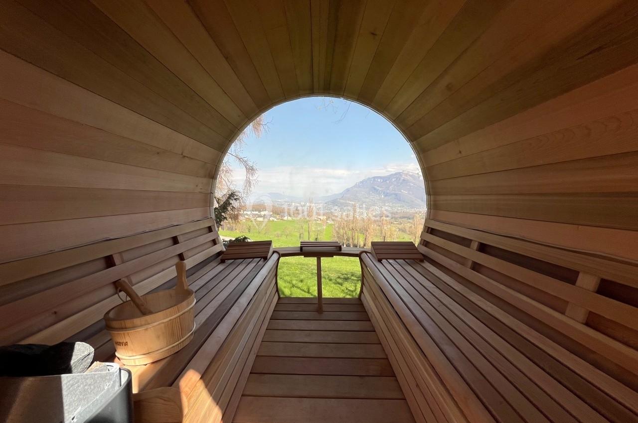 Intérieur d'un sauna en bois en forme de tonneau avec vue sur un paysage verdoyant et des montagnes.