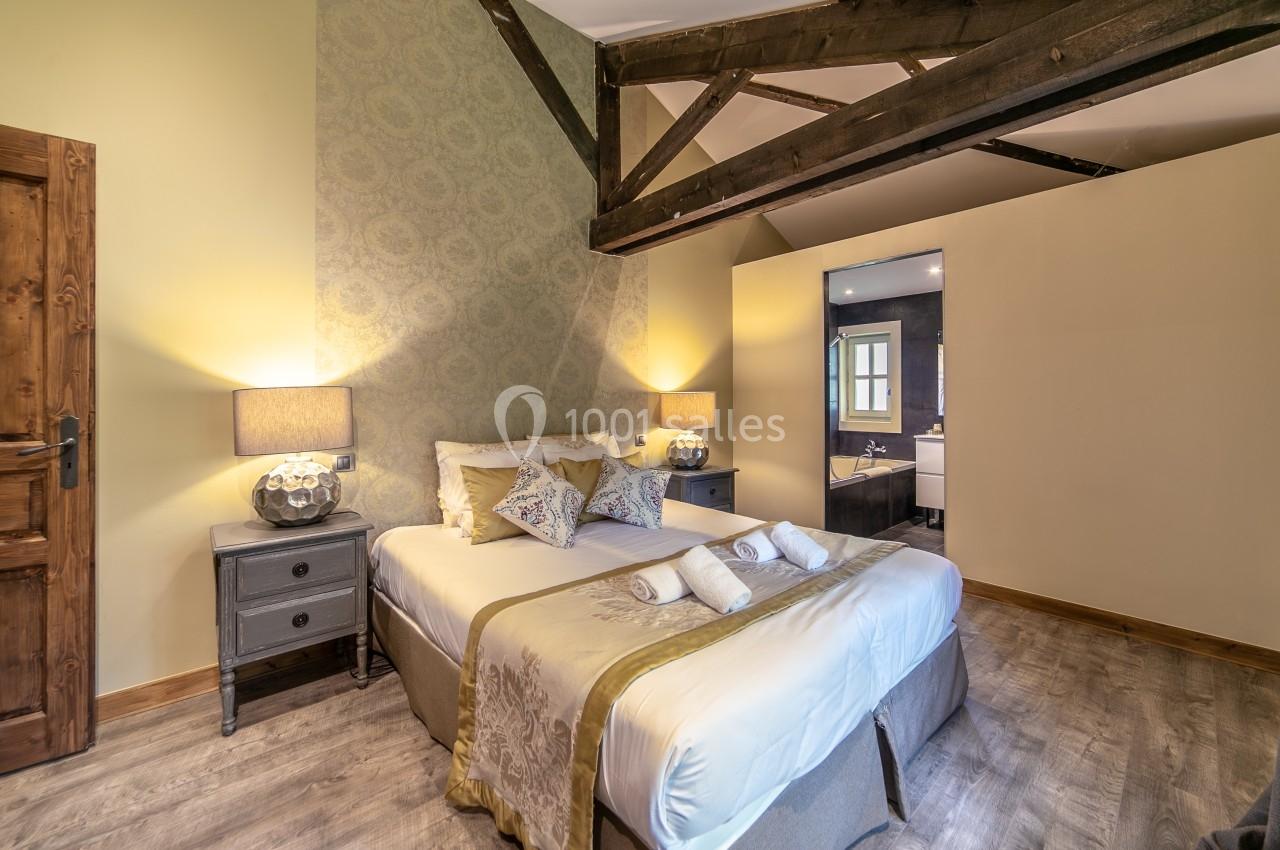 Chambre lumineuse avec lit double, poutres apparentes, tables de chevet et vue partielle sur une salle de bain moderne.