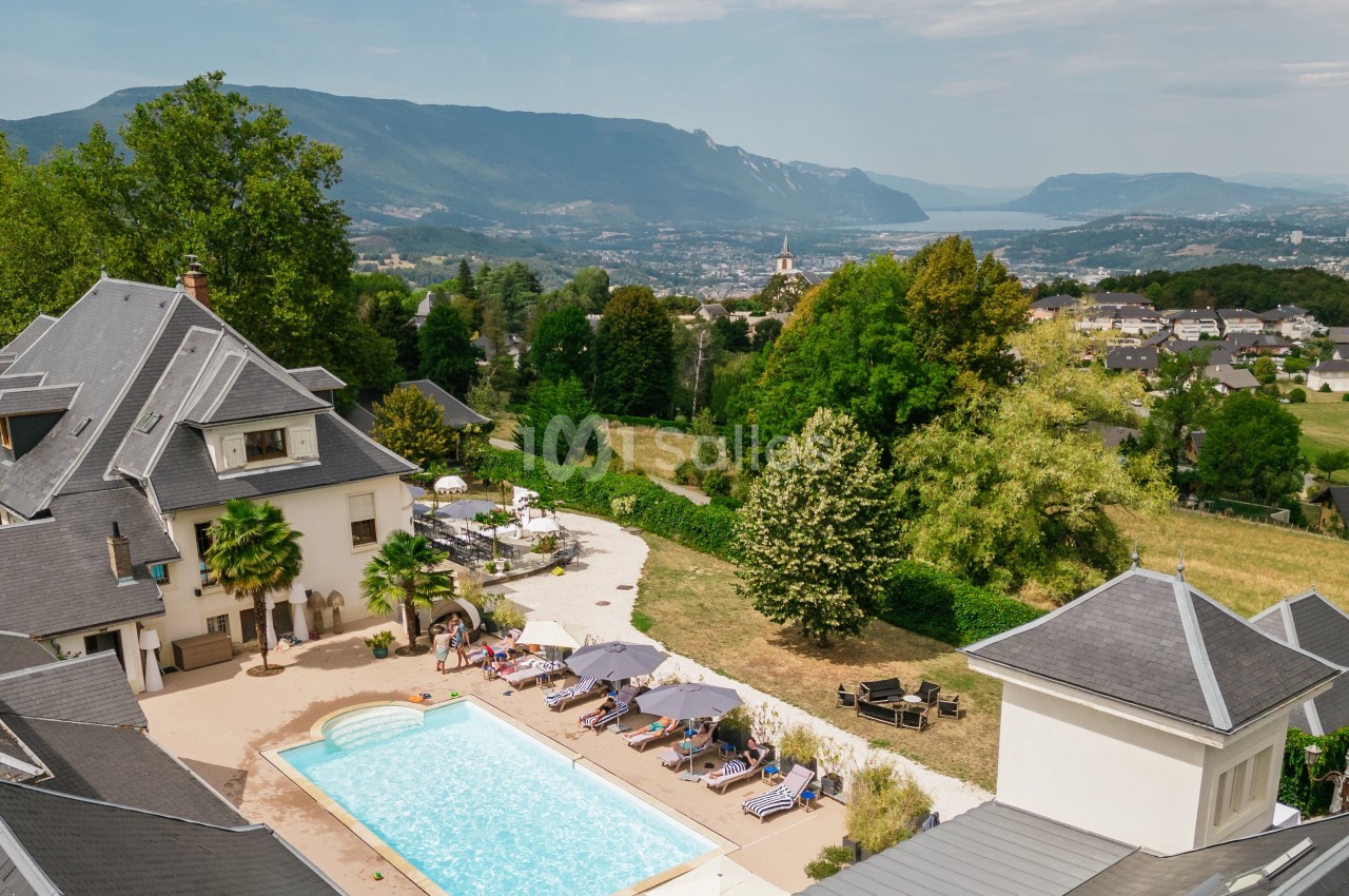 Vue aérienne d'une maison avec piscine entourée de verdure, offrant un panorama sur des montagnes et un lac au loin.
