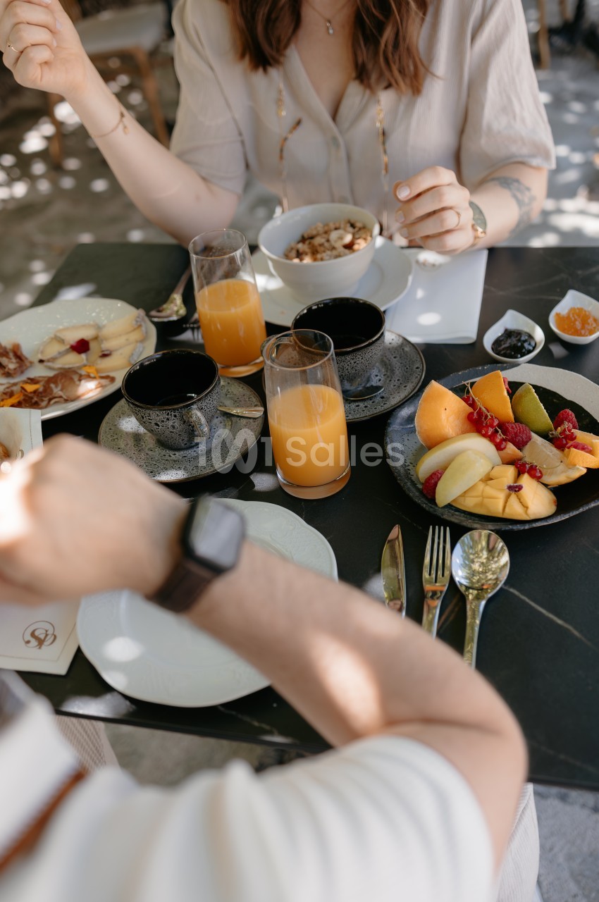 Deux personnes prennent un petit-déjeuner avec fruits, jus d'orange, céréales et couverts sur une table noire.