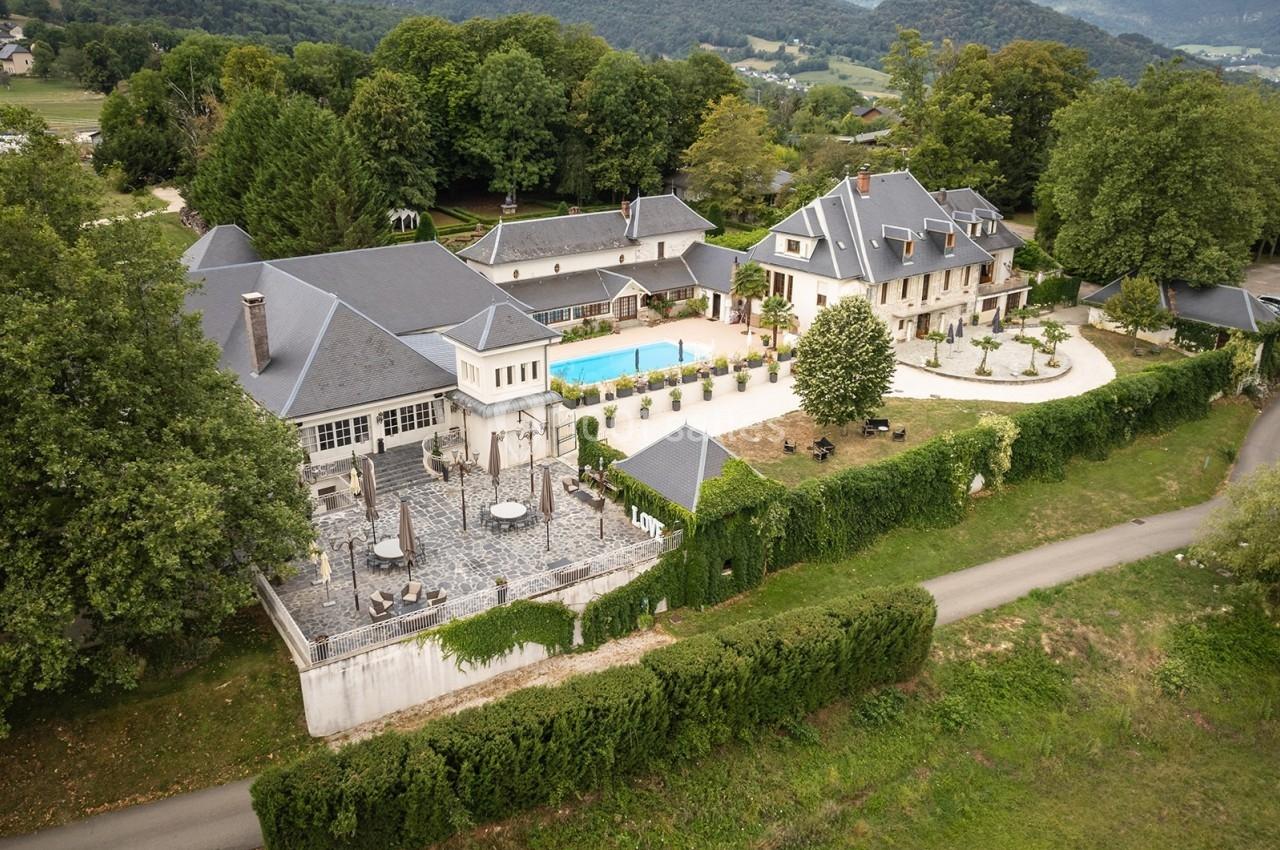 Vue aérienne d'un domaine avec bâtiments élégants, piscine centrale, terrasse et jardin, entouré de verdure.