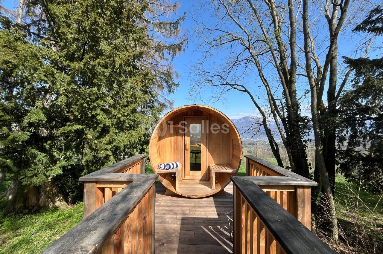 Cabane en bois en forme de tonneau avec terrasse, entourée d'arbres et offrant une vue dégagée sur un paysage vallonné.