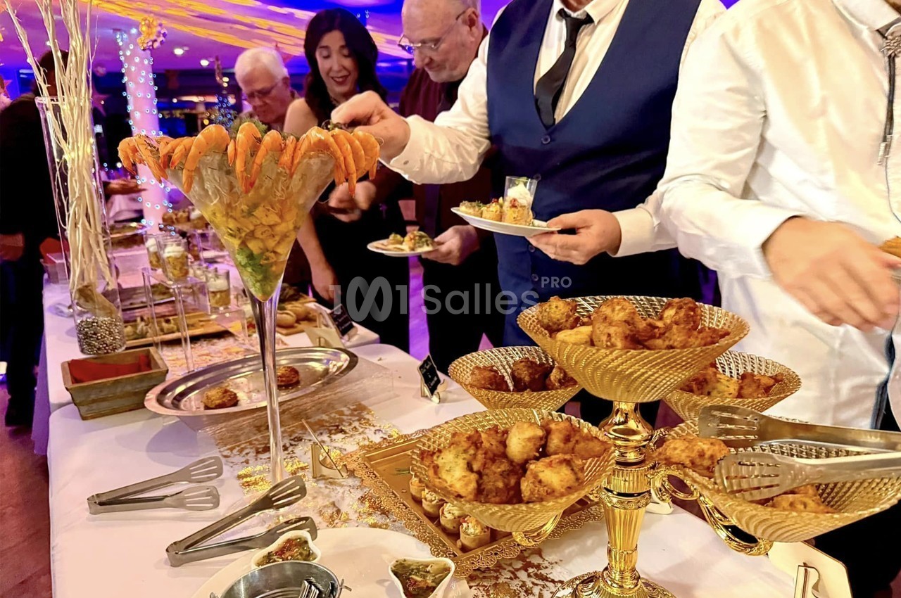 Buffet élégant avec verrine de crevettes et plats dorés, des convives se servant dans une ambiance festive.