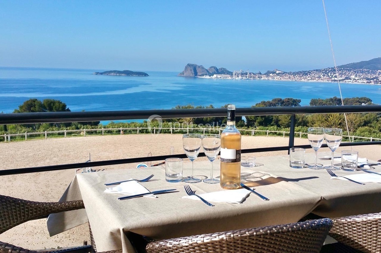 Table dressée avec une bouteille de vin, verres et couverts, offrant une vue panoramique sur la mer et les collines.