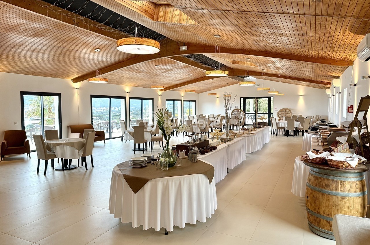 Salle lumineuse avec tables dressées, buffet central, plafond en bois et grandes baies vitrées donnant sur l'extérieur.