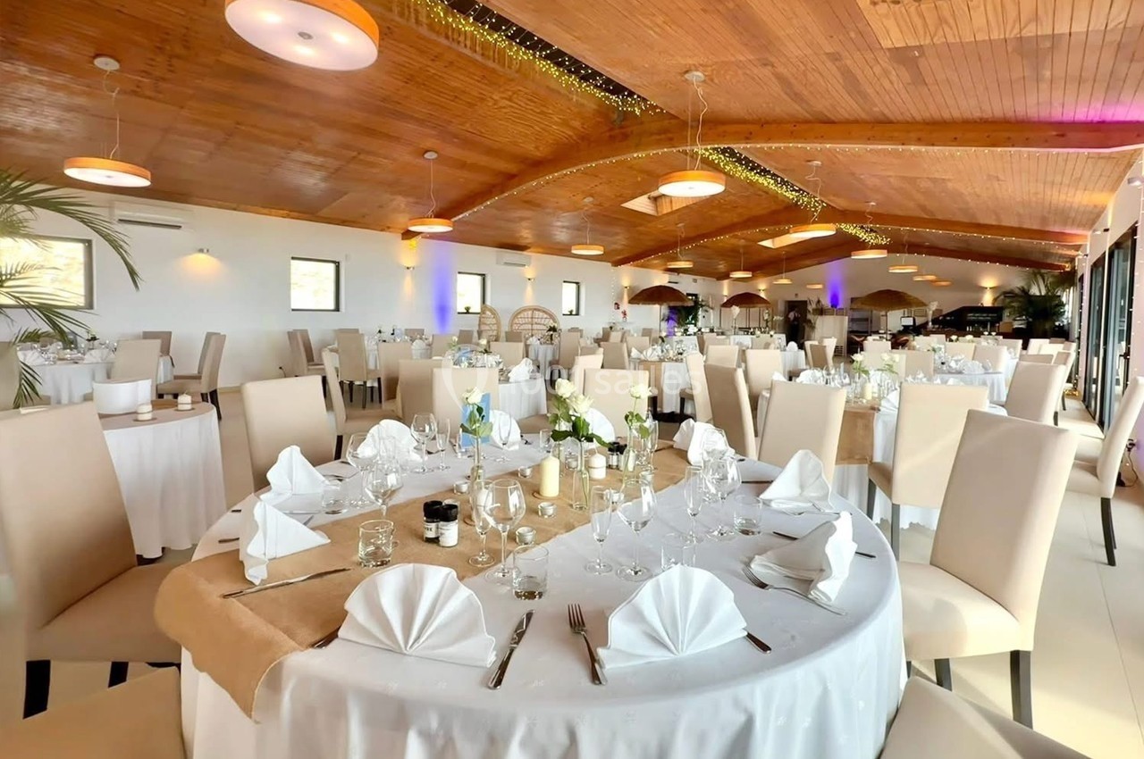 Salle de réception lumineuse avec tables rondes dressées, nappes blanches et décoration élégante sous un plafond en bois.