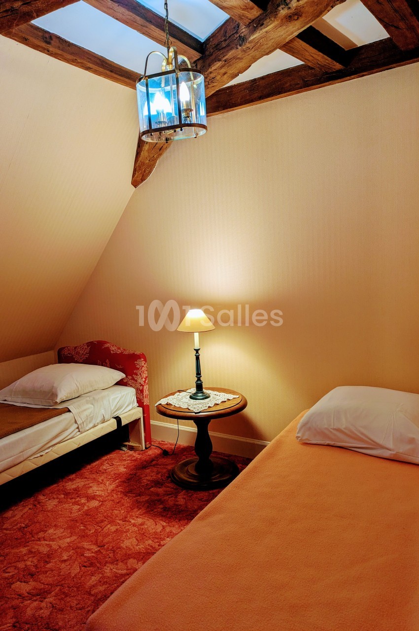 Deux lits simples dans une chambre mansardée avec poutres apparentes, éclairée par une lampe de chevet et un lustre.