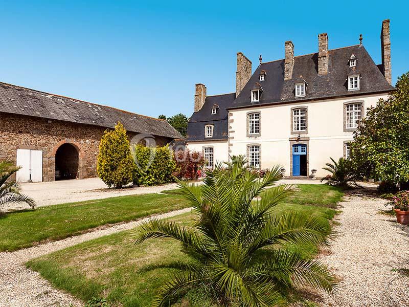 Façade d'un manoir ancien en pierre avec toit en ardoise, entouré d'un jardin arboré et d'une cour gravillonnée.