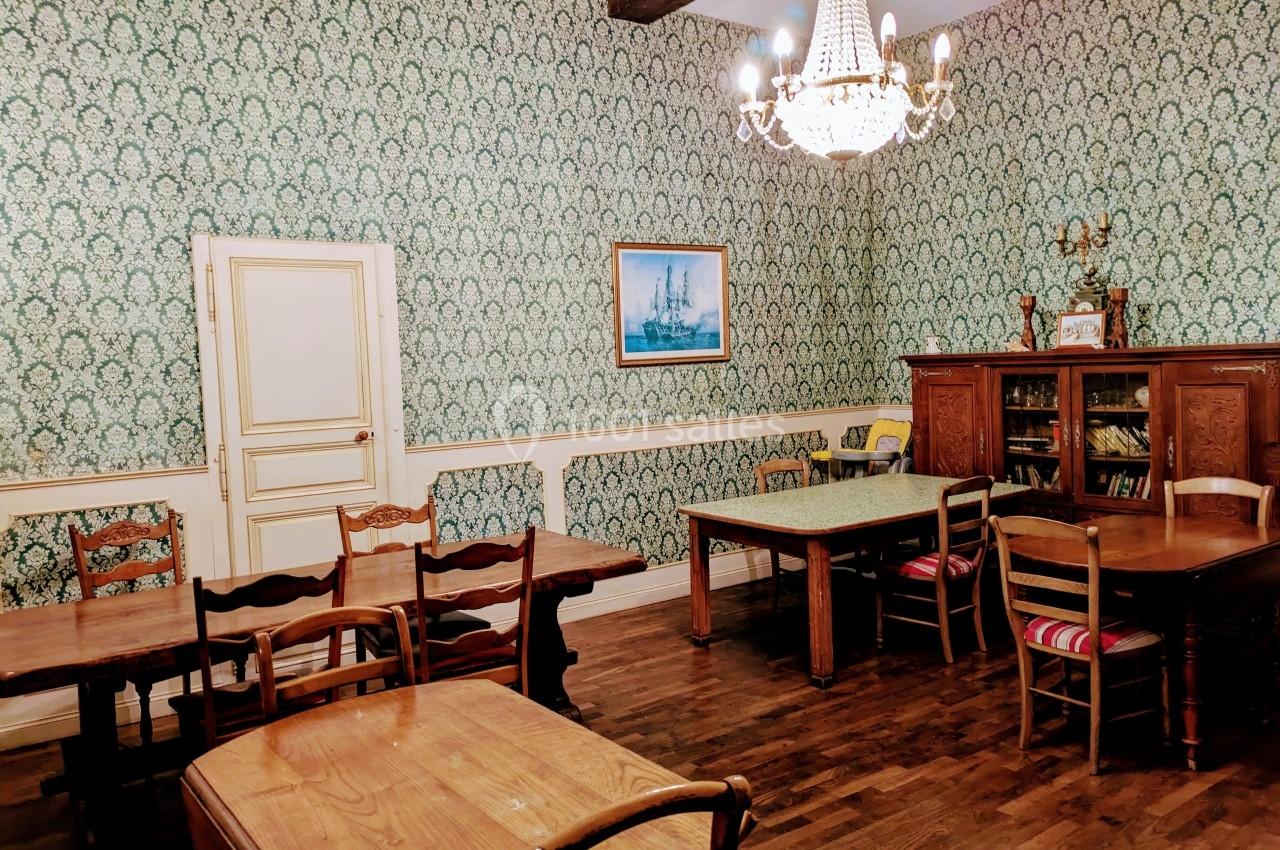 Salle à manger avec tables en bois, chaises assorties, papier peint à motifs verts, lustre et meuble bibliothèque.
