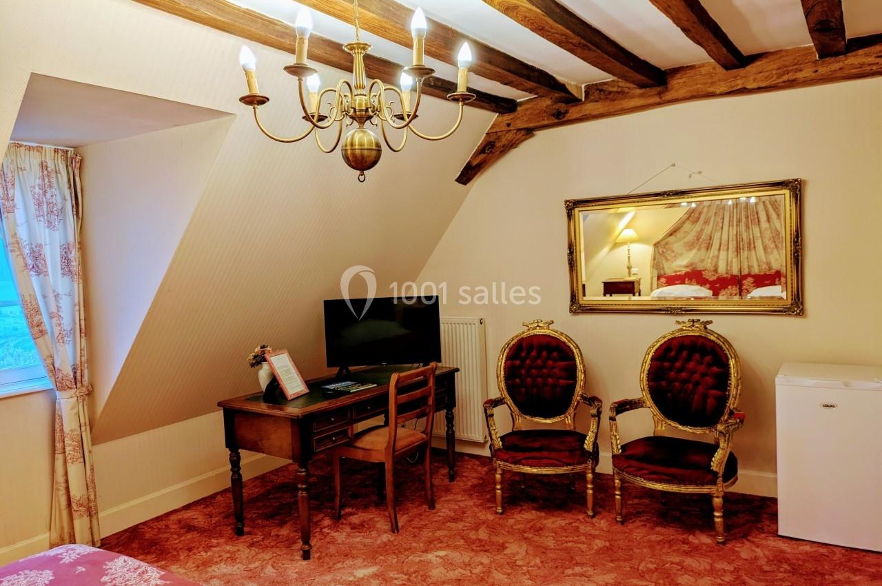 Chambre avec poutres apparentes, bureau, fauteuils anciens, téléviseur, mini-frigo et miroir mural.