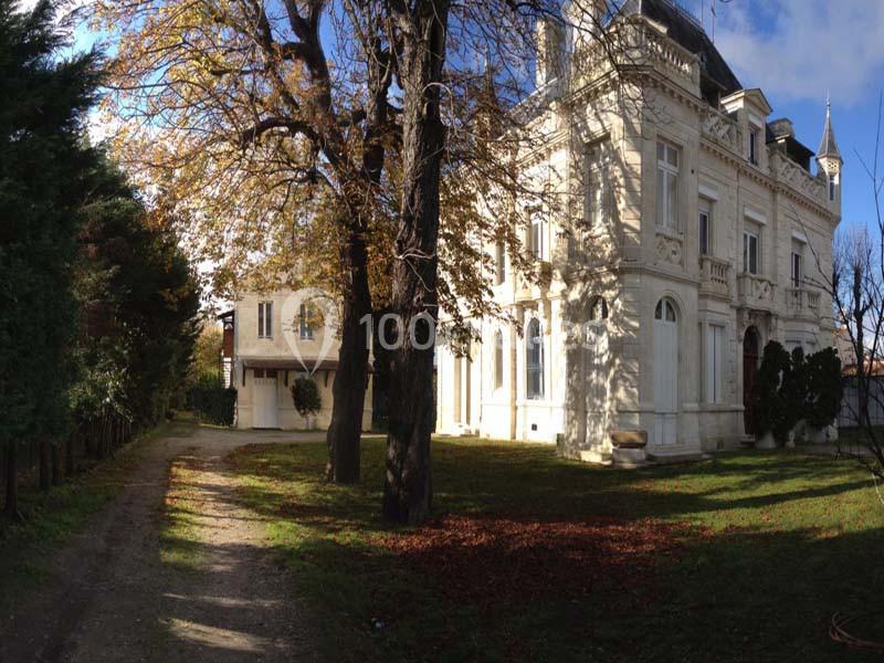 Grande maison en pierre blanche entourée d'arbres, avec une allée et un jardin ensoleillé à l'avant.