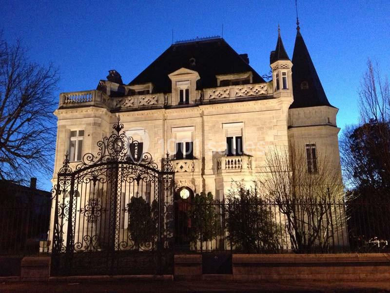 Façade d'un manoir éclairé en soirée, avec une grille en fer forgé au premier plan et un ciel bleu foncé en arrière-plan.