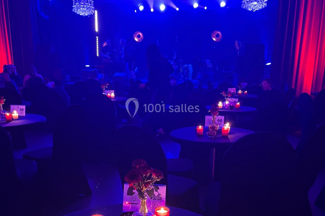 Salle sombre avec tables éclairées par des bougies rouges, scène illuminée en arrière-plan sous des projecteurs bleus.