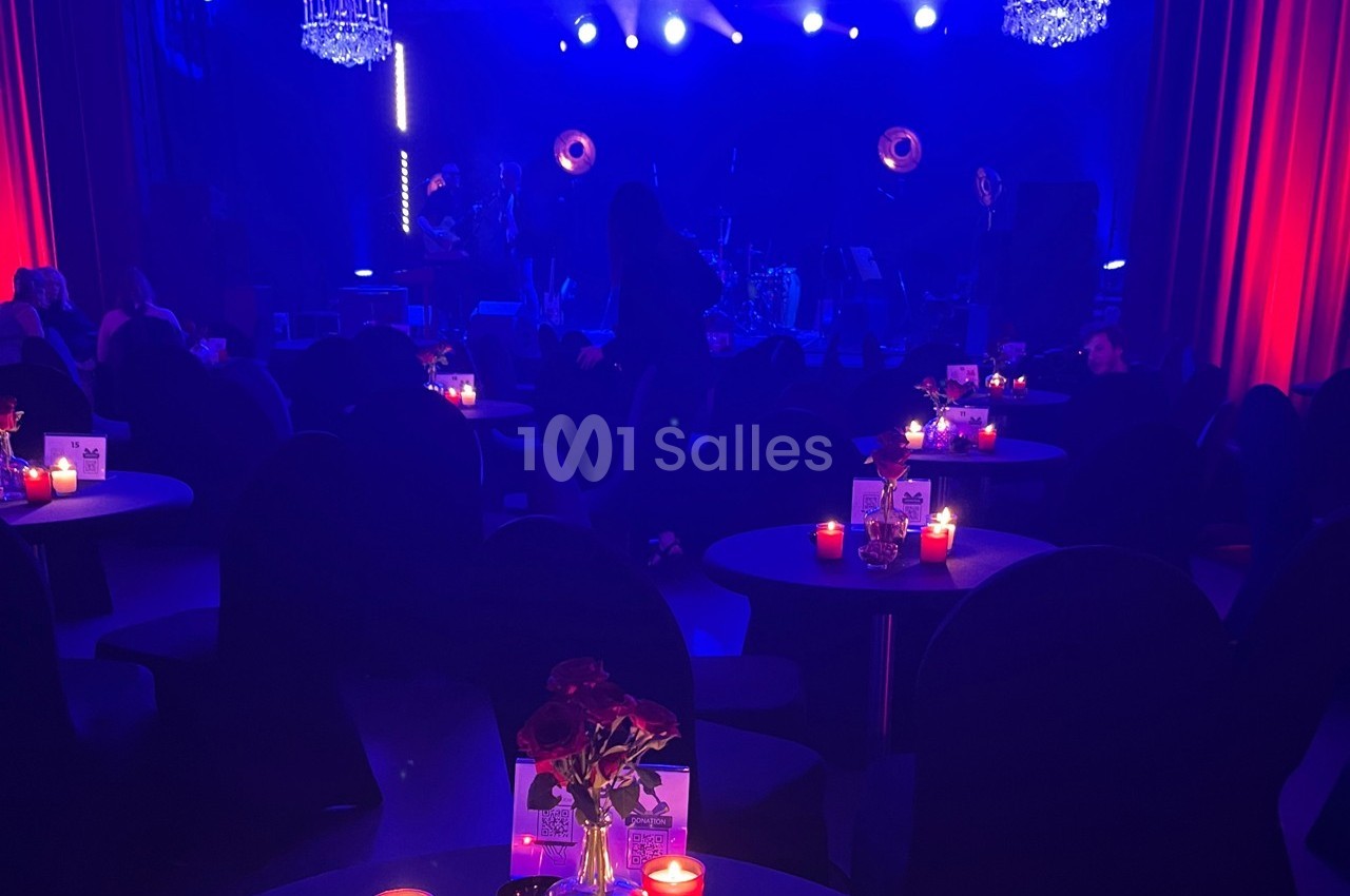 Salle sombre avec tables éclairées par des bougies rouges, scène illuminée en arrière-plan sous des projecteurs bleus.