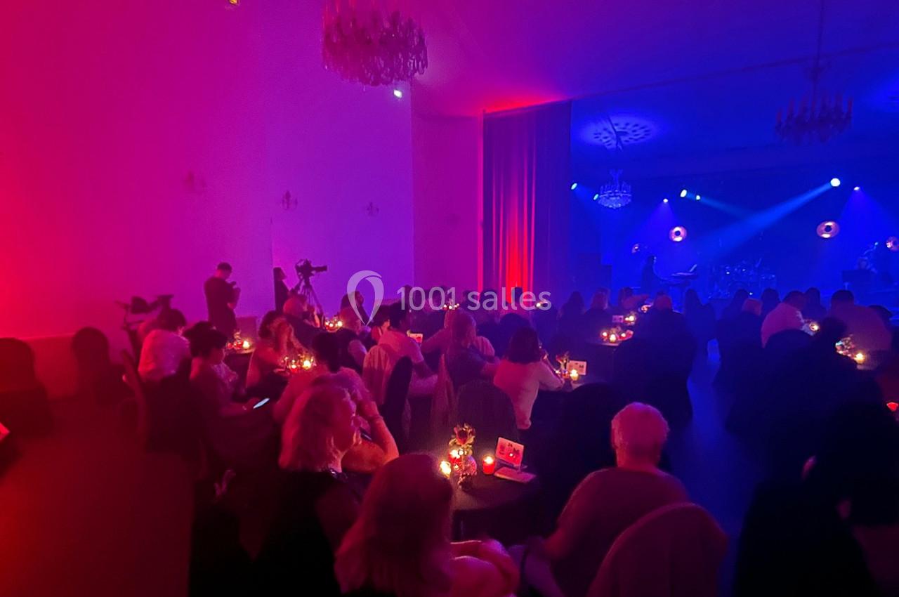 Salle tamisée avec des spectateurs assis à des tables éclairées par des bougies, face à une scène illuminée en rouge et bleu.