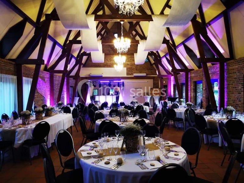 Location salle Watten (Nord) - Le Domaine du Fil de l’Eau #2 Salle de réception décorée avec des tables rondes dressées, éclairage chaleureux et poutres apparentes en bois.