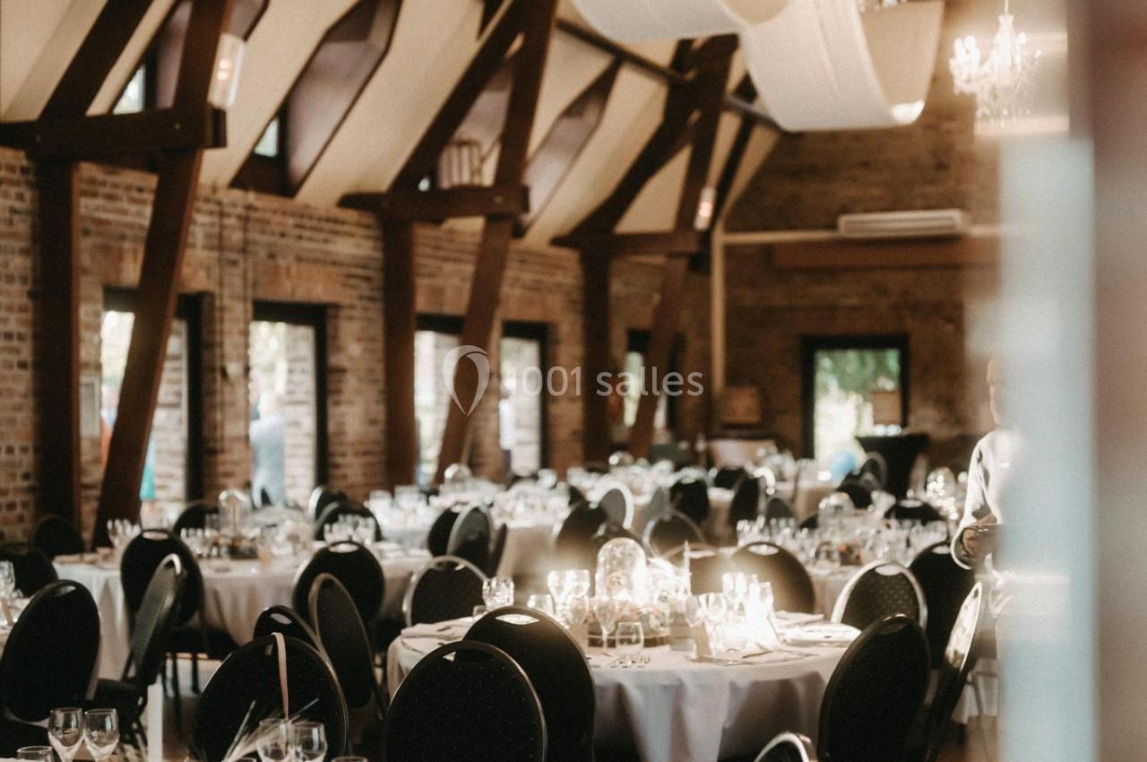 Location salle Watten (Nord) - Le Domaine du Fil de l’Eau #20 Salle de réception élégante avec tables rondes dressées, nappes blanches, chandeliers et poutres apparentes en bois.