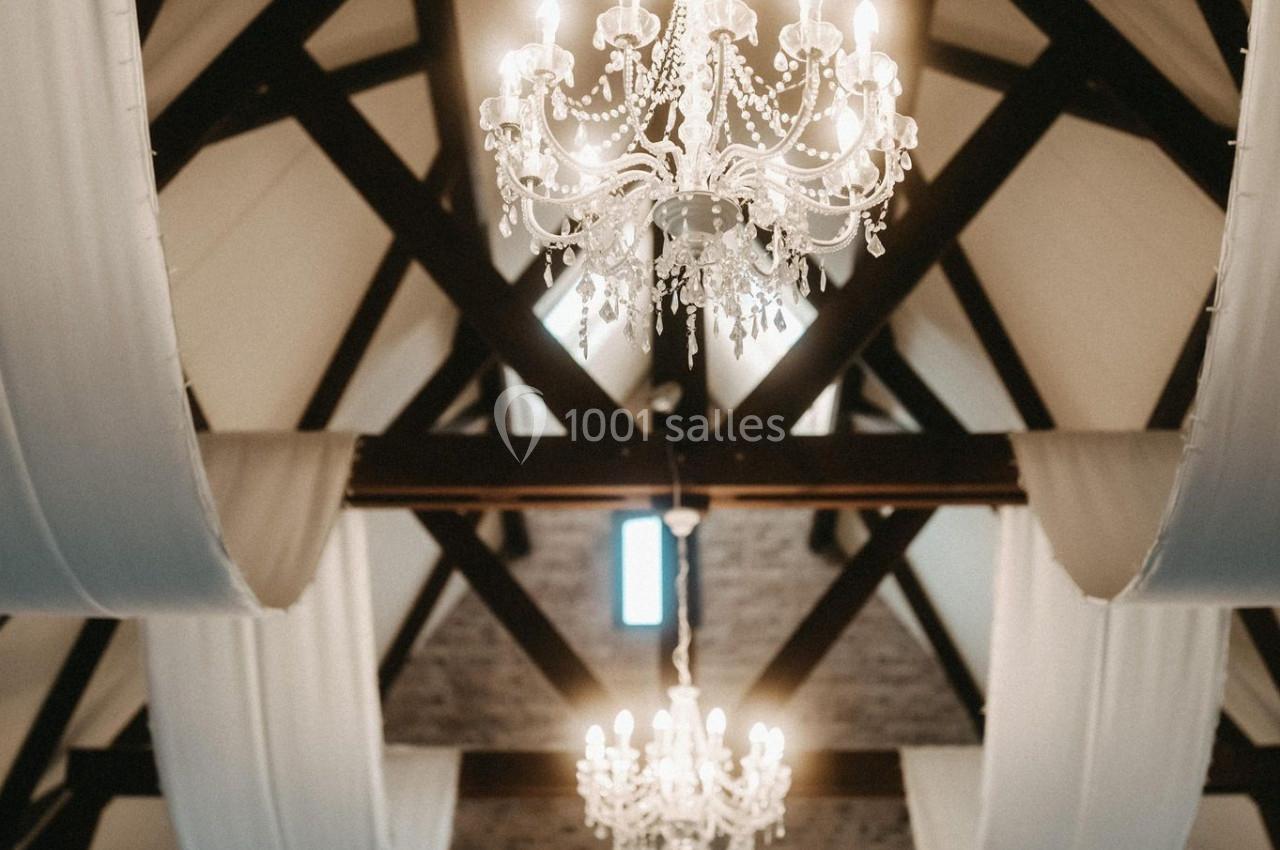 Location salle Watten (Nord) - Le Domaine du Fil de l’Eau #9 Plafond avec poutres apparentes, lustres en cristal allumés et drapés blancs suspendus dans une salle élégante.