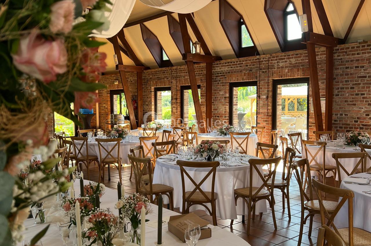 Location salle Watten (Nord) - Le Domaine du Fil de l’Eau #5 Salle de réception lumineuse avec tables rondes décorées de fleurs, nappes blanches et chaises en bois.