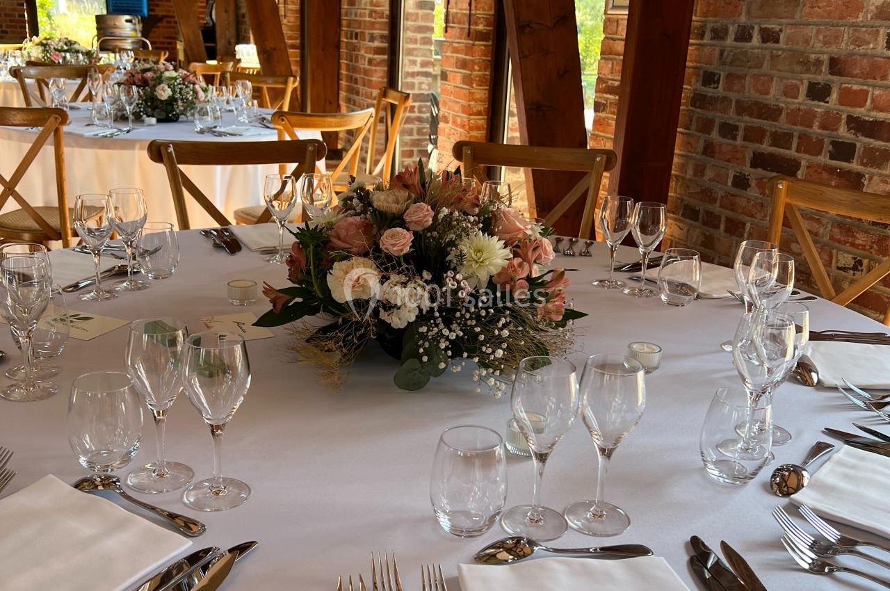 Location salle Watten (Nord) - Le Domaine du Fil de l’Eau #6 Table ronde dressée avec nappes blanches, couverts élégants et centre de table floral dans une salle en briques apparentes.