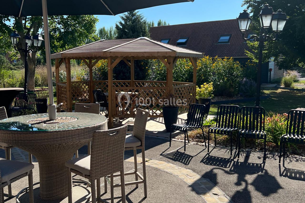 Location salle Watten (Nord) - Le Domaine du Fil de l’Eau #11 Terrasse ensoleillée avec table, chaises, parasol et pergola en bois dans un jardin verdoyant.
