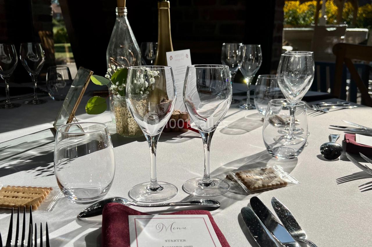 Location salle Watten (Nord) - Le Domaine du Fil de l’Eau #24 Table élégamment dressée avec menu, couverts, verres et une fleur rouge en centre de table, éclairée par la lumière…