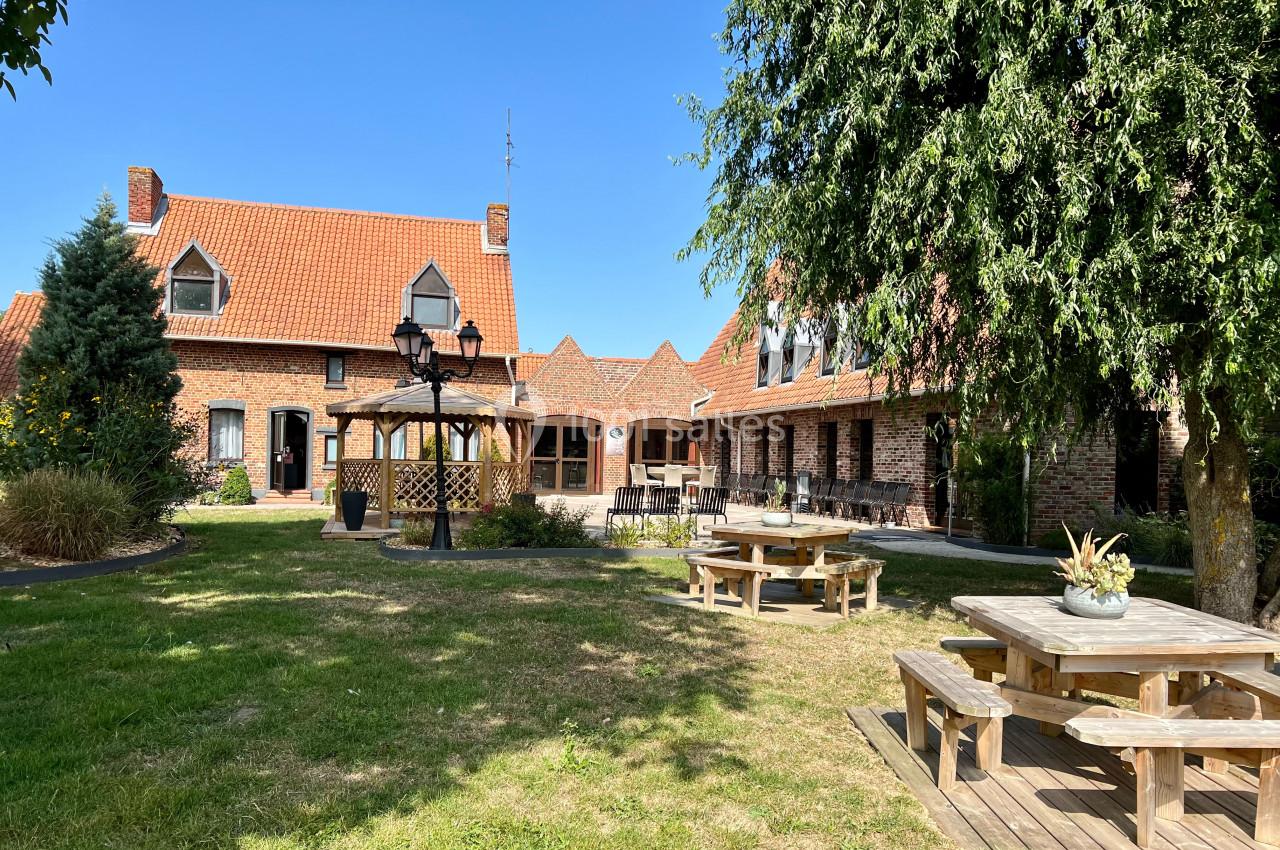 Location salle Watten (Nord) - Le Domaine du Fil de l’Eau #23 Cour avec jardin, tables en bois, kiosque et bâtiments en briques rouges sous un ciel bleu.