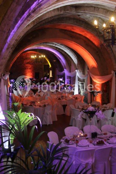Salle voûtée décorée pour un événement, avec tables dressées, nappes blanches et éclairage coloré.