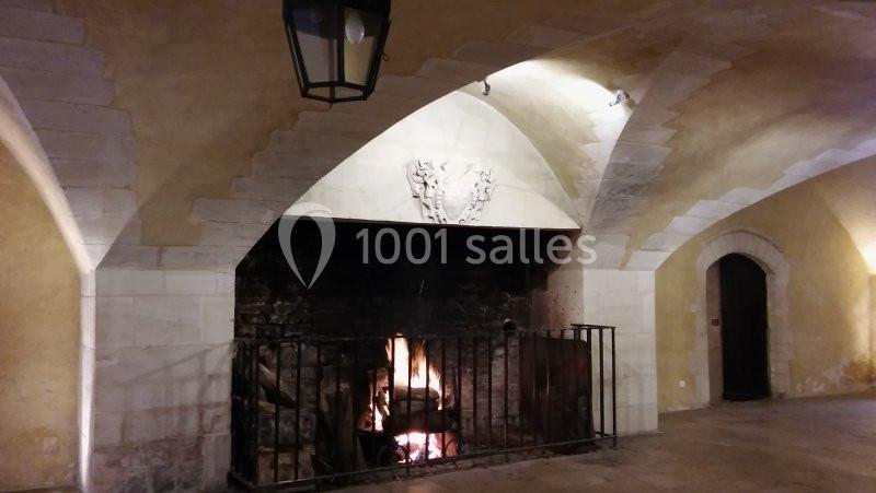 Cheminée en pierre avec feu de bois allumé, entourée d'une grille, dans une pièce voûtée aux murs clairs.