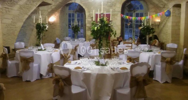 Salle voûtée décorée pour un événement, avec tables rondes dressées, nappes blanches et chaises ornées de rubans.