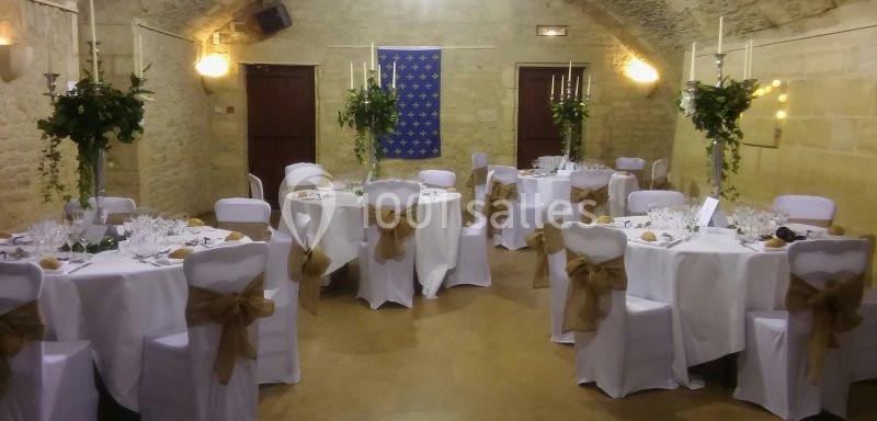 Salle en pierre décorée pour un événement, avec tables rondes dressées, chaises blanches et centres de table fleuris.