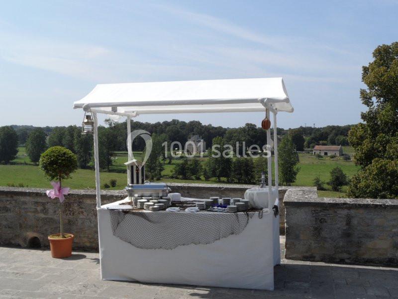 Stand extérieur avec nappe blanche, vaisselle et décorations, situé sur une terrasse surplombant un paysage verdoyant.