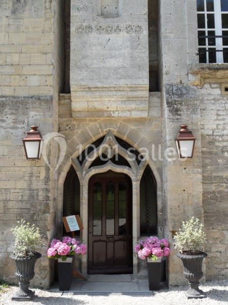 Entrée d'un bâtiment en pierre avec une porte en bois, encadrée par des arches et des pots de fleurs roses.